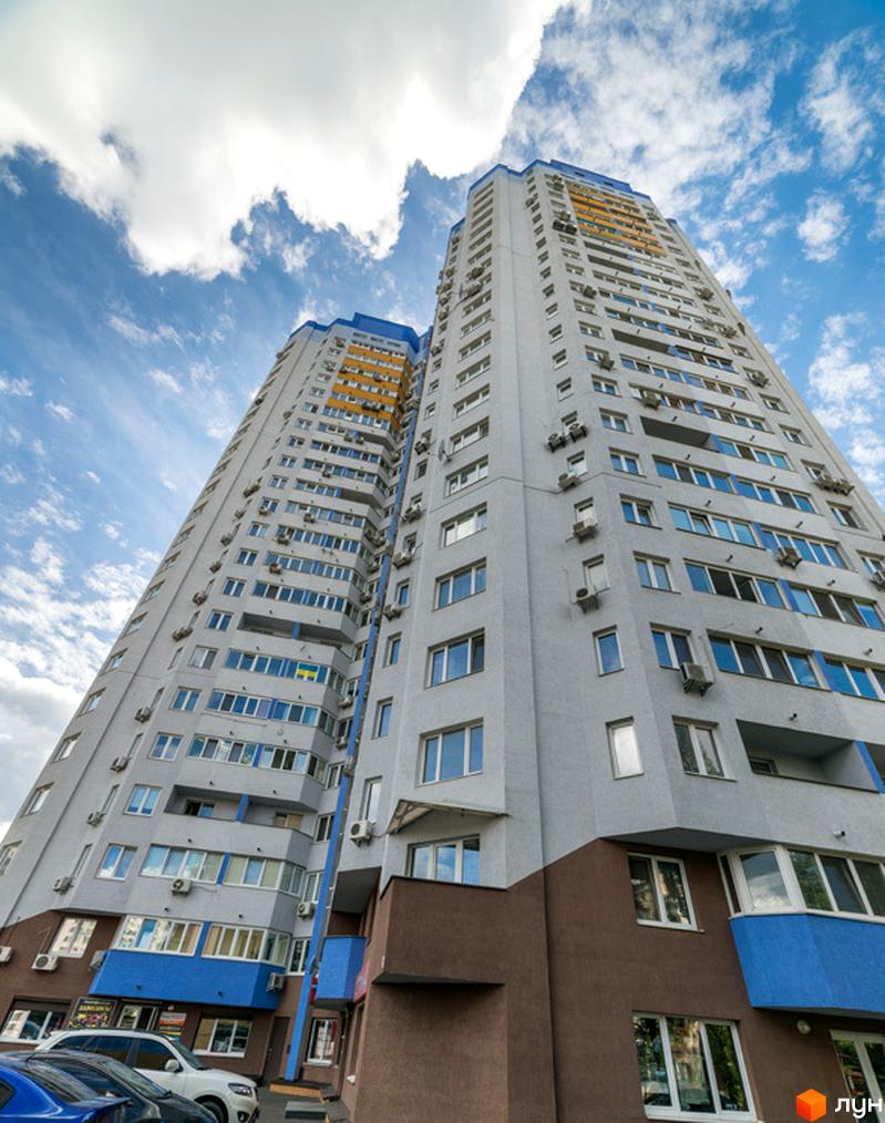 Високі житлові будинки Житлового комплексу Авіатор світло-сірого кольору з синіми та жовтими акцентами. На прибудинковій території припарковані автомобілі.