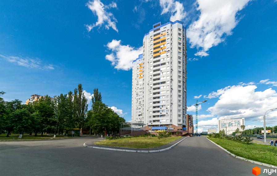 Багатоповерховий житловий комплекс Авіатор світло-сірого кольору з синіми та помаранчевими елементами. Перед будинком зелені насадження та асфальтована дорога.