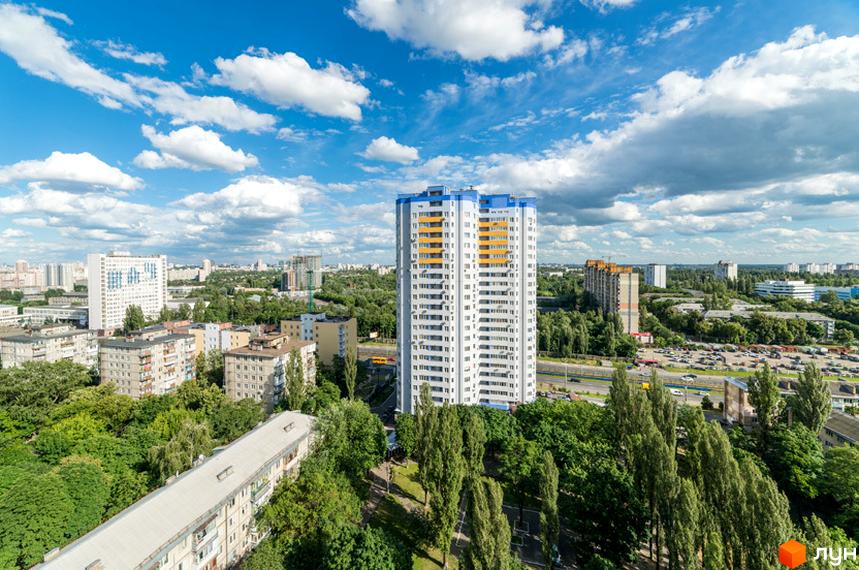 Багатоповерховий будинок Житловий комплекс Авіатор білого кольору з синіми та жовтими елементами. Навколо багато зелених дерев, інші житлові будинки та міський пейзаж.