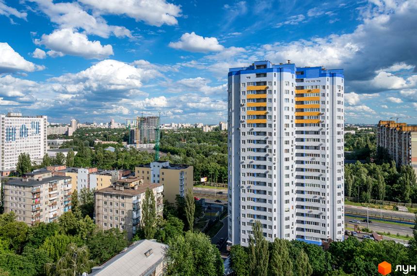 Житловий комплекс Авіатор з високими світло-сірими фасадами та синьо-жовтими елементами. Навколо багато зелених насаджень та міська забудова.