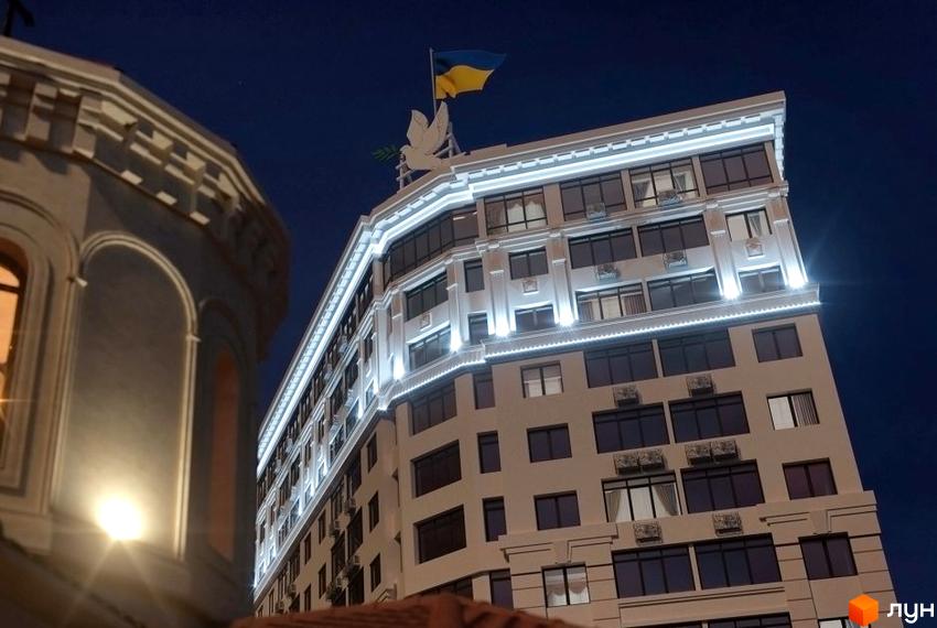 Зображення фасаду житлового комплексу Тридцять перша перлина з українським прапором на даху. Вигляд багатоповерхового будинку вночі.
