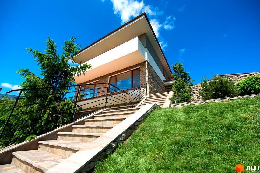 Двоповерховий будинок з панорамними вікнами та кам'яним фасадом. Перед будинком розташовані сходи та зелена галявина з рослинністю.