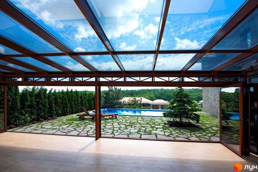 Критий басейн з прозорим дахом та розсувними скляними стінами. На території є газон з камінням, шезлонги та дерева, а також вид на басейн та котеджі.