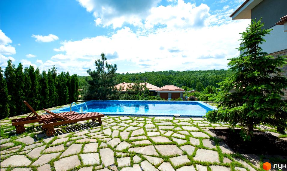 На фото зображено будинок з басейном на території котеджного містечка. Навколо басейну викладено кам'яну плитку з травою, а також є шезлонги для відпочинку.