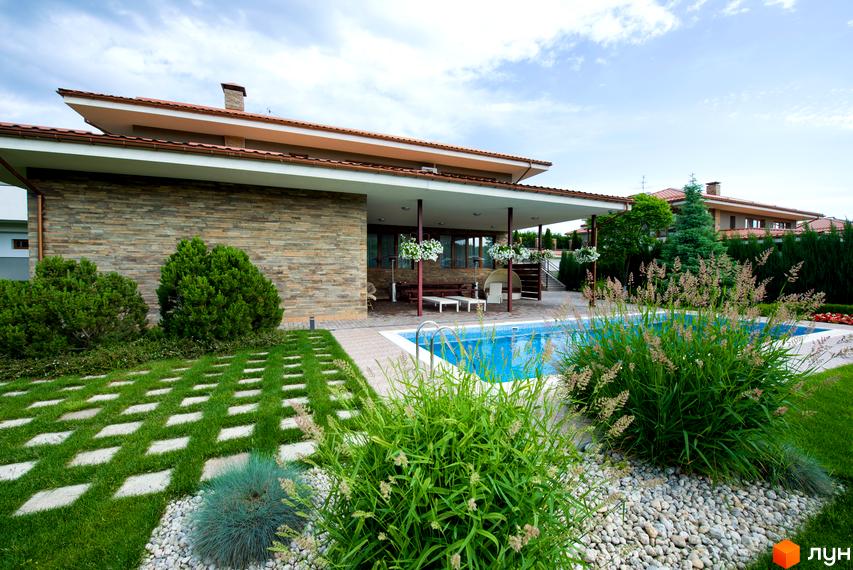 Будинок з кам'яним фасадом та терасою біля басейну. На території газон з доріжкою з плитки та декоративні рослини.