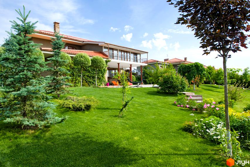 Двоповерховий будинок з червоною черепицею та балконом. Простора зелена прибудинкова територія з газоном, деревами та квітами.