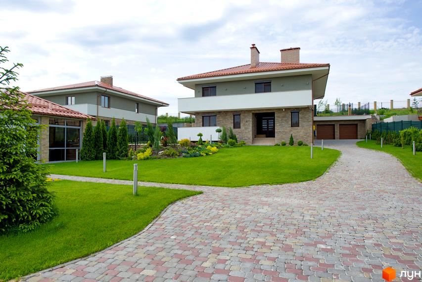 Двоповерховий будинок з черепичним дахом та кам'яним фасадом. На прибудинковій території газон, доріжки з бруківки та декоративні рослини.