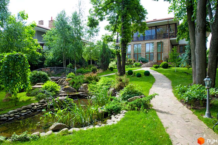 Будинок з кам'яним фасадом та великими вікнами. На прибудинковій території доглянутий газон, доріжка з каменю та декоративний ставок з містком.