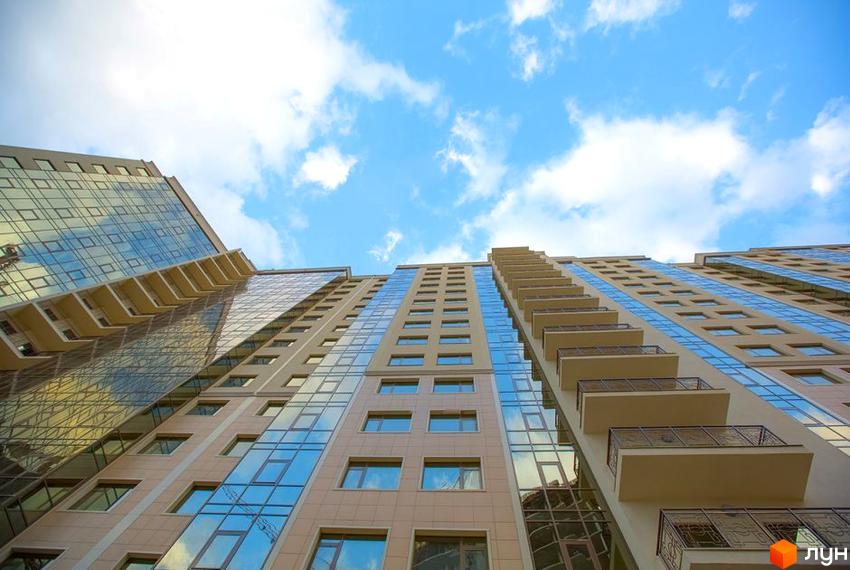 Зовнішній вигляд житлового комплексу Четвертая Жемчужина, що складається з висотних будинків з панорамним склінням та балконами. Фото новобудови з низького ракурсу на тлі блакитного неба з хмарами.