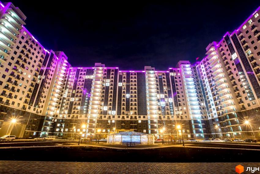 Житловий комплекс Пятая Жемчужина вночі з фіолетовою підсвіткою. Зовнішній вигляд будинку з багатьма вікнами та балконами, прибудинкова територія з освітленням та автомобілями.