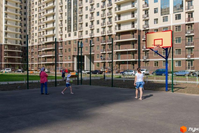 Житловий комплекс Пятая Жемчужина з баскетбольним майданчиком та людьми на території. Сучасна архітектура та зелена зона біля будинку.