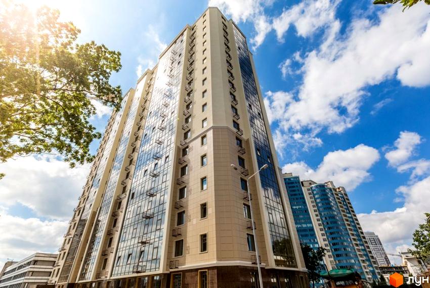 Високий будинок ЖК Шоста Перлина з великими вікнами на фоні блакитного неба з хмарами. Поруч зелені дерева та інші будівлі.