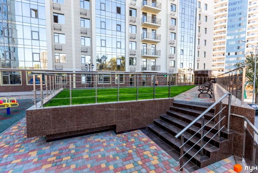 Зображення фасаду багатоповерхового будинку ЖК Шоста Перлина з скляними вікнами. Перед будинком розташована прибудинкова територія з газоном, сходами та кованими лавками.