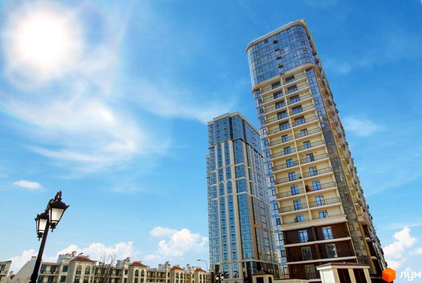 Високі будинки з скляними фасадами на тлі блакитного неба. Поруч ліхтарі та будівлі з червоними дахами.