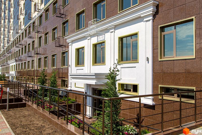 Фасад Житлового комплексу Двадцята Перлина з коричневої плитки та білими декоративними елементами. Перед будинком пандус з металевими поручнями та зелені насадження.