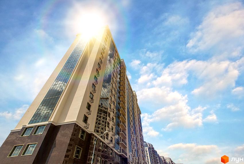 Житловий комплекс Двадцять перша Перлина: високий багатоповерховий будинок з світлими стінами та скляними фасадами, освітлений сонцем на тлі блакитного неба з хмарами.