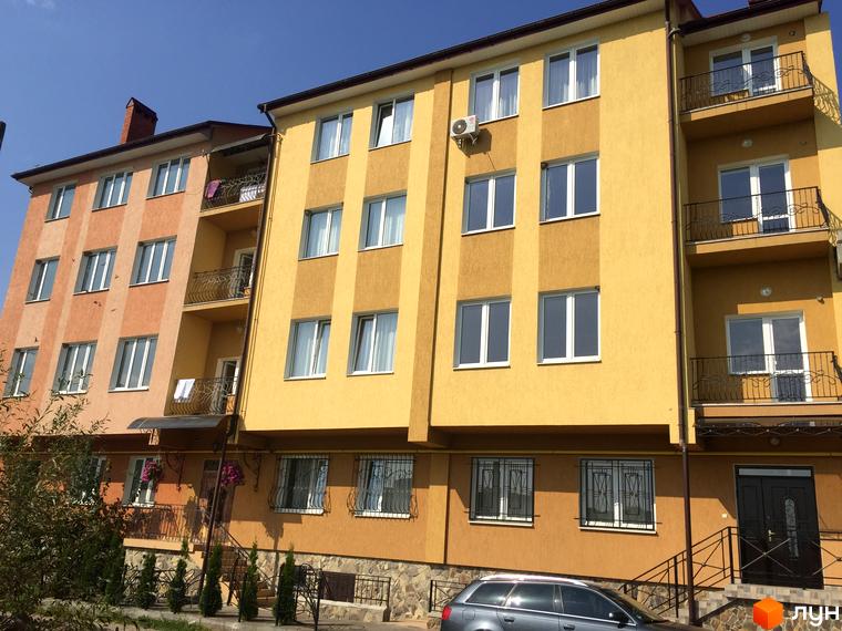 Фасад жовтого та помаранчевого будинку з балконами та вікнами. На прибудинковій території видно зелені насадження та автомобіль.