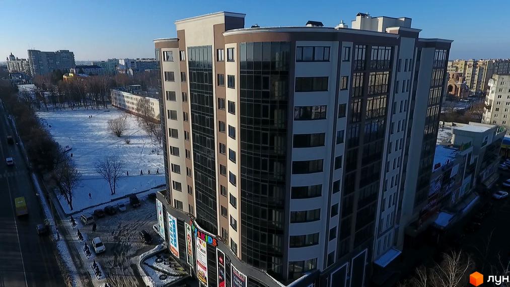 Зовнішній вигляд новобудови ЖК Арена з панорамними вікнами та сірими елементами фасаду. Навколо будівлі розташована прибудинкова територія з деревами та припаркованими автомобілями.