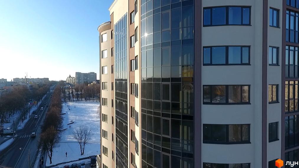 Зовнішній вигляд новобудови ЖК Арена з панорамними вікнами та міським пейзажем. Навколо будинку розташована засніжена територія та дорога з автомобілями.