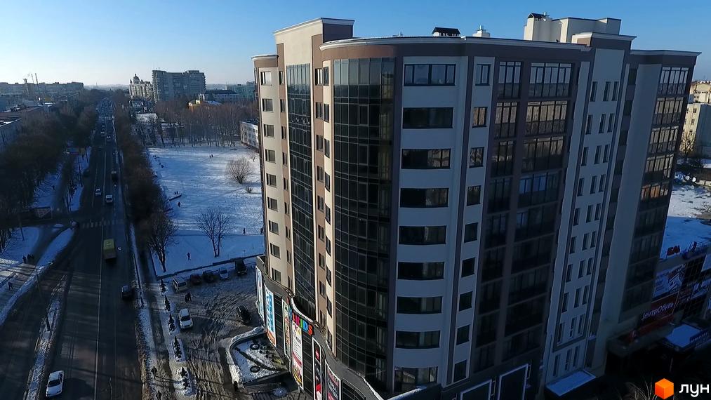 Зовнішній вигляд новобудови ЖК Арена з панорамними вікнами та сірими елементами фасаду. Навколо будівлі розташована дорога з автомобілями та пішохідна зона.