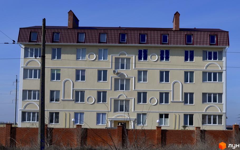 Зовнішній вигляд багатоповерхового будинку ЖК Зелений гай з червоним дахом та світлими стінами. Навколо будинку видно цегляний паркан та дерева.