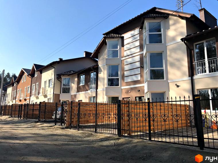 Котеджне містечко КМ Сонячний 2. Фасади будинків у світлих тонах, з коричневими та бежевими елементами, з балконами та великими вікнами. Огороджена територія з дерев'яним парканом та кованими елементами.