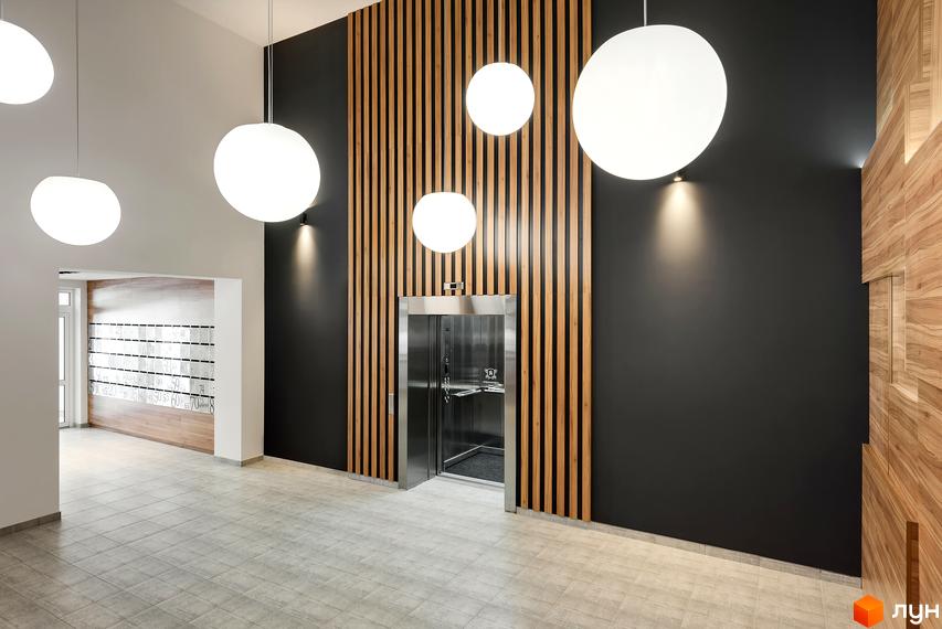 Холл житлового комплексу Continent з ліфтом, оздоблений деревом та чорною фарбою. Яскраве освітлення та світла плитка на підлозі створюють відчуття простору.