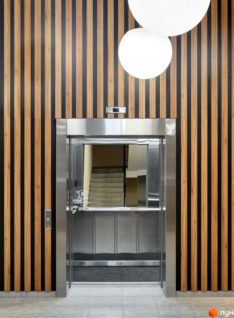Ліфт в холі ЖК Continent з оздобленням з дерева та темних смуг. На стіні розташовані круглі світильники.