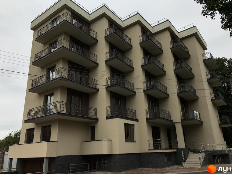 Зображення фасаду багатоповерхового будинку. На фасаді будинку розташовані балкони з кованими огорожами. Біля будинку є сходи та невелика прибудинкова територія.