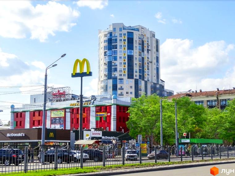 Житловий комплекс Аксіома - висотна будівля з блакитними та білими вікнами. Поруч розташований Макдональдс та зелені дерева.