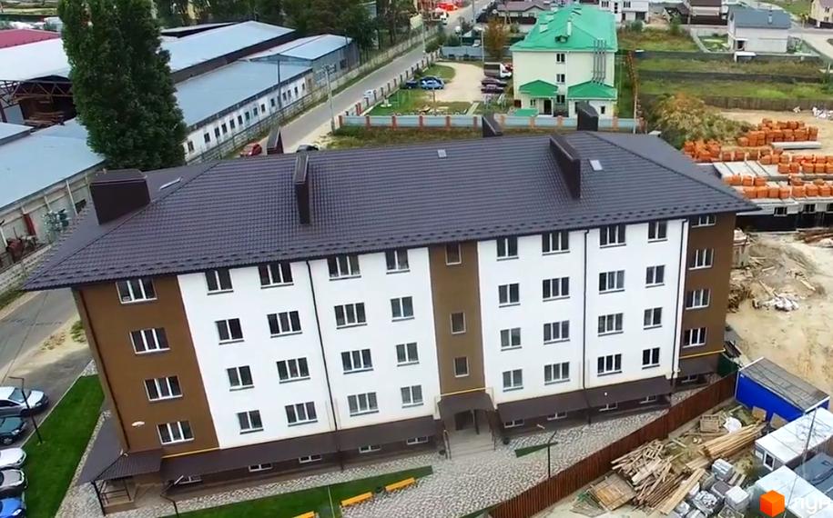 Зовнішній вигляд багатоквартирного будинку ЖК Оксамитовий з коричнево-білим фасадом та темним дахом. На прибудинковій території видно паркування, зелену зону та будівельні матеріали.