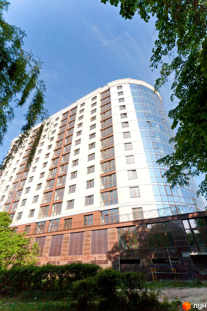 Багатоповерховий будинок Житлового комплексу Paradise з білим фасадом та великими вікнами, включаючи заокруглену скляну частину. Оточений зеленою рослинністю під блакитним небом.