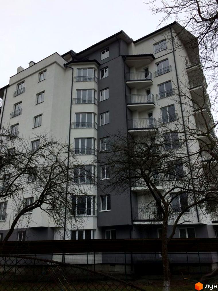 Зовнішній вигляд новобудови ЖК Гармонія з білими та сірими фасадами, балконами та вікнами. Навколо будівлі ростуть дерева без листя.