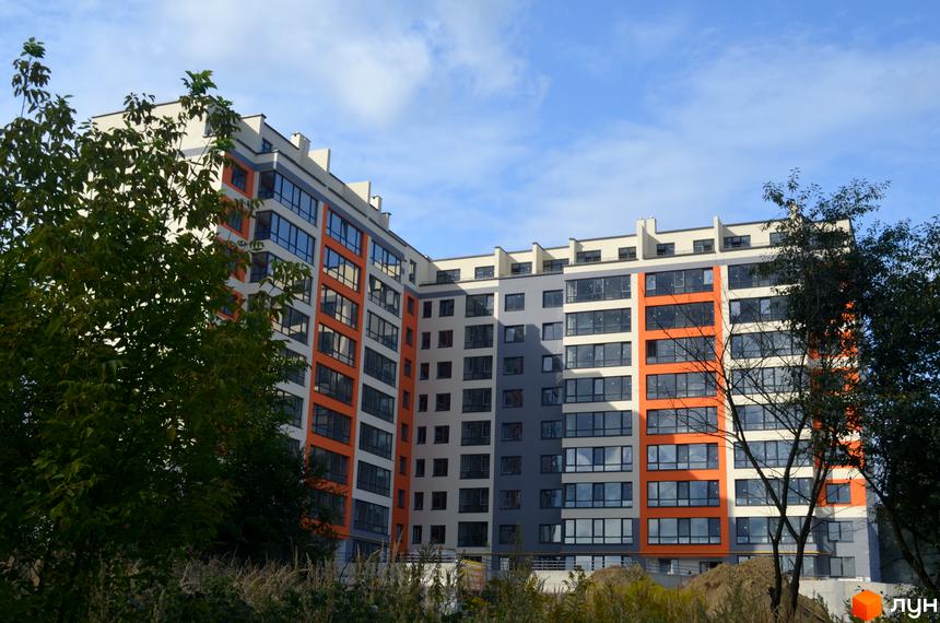 Житловий комплекс Deluxe: багатоповерховий будинок з помаранчевими, сірими та білими фасадами. Навколо видно зелені дерева та траву.