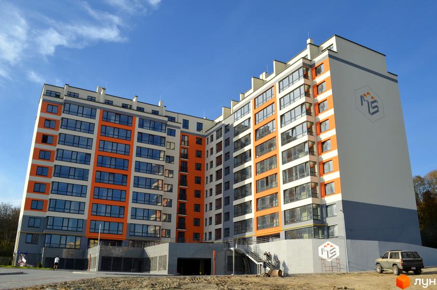 Багатоповерховий житловий комплекс Deluxe з біло-помаранчевим фасадом та великими вікнами. Навколо будинку видно ділянку з ґрунтом та під'їзні шляхи.