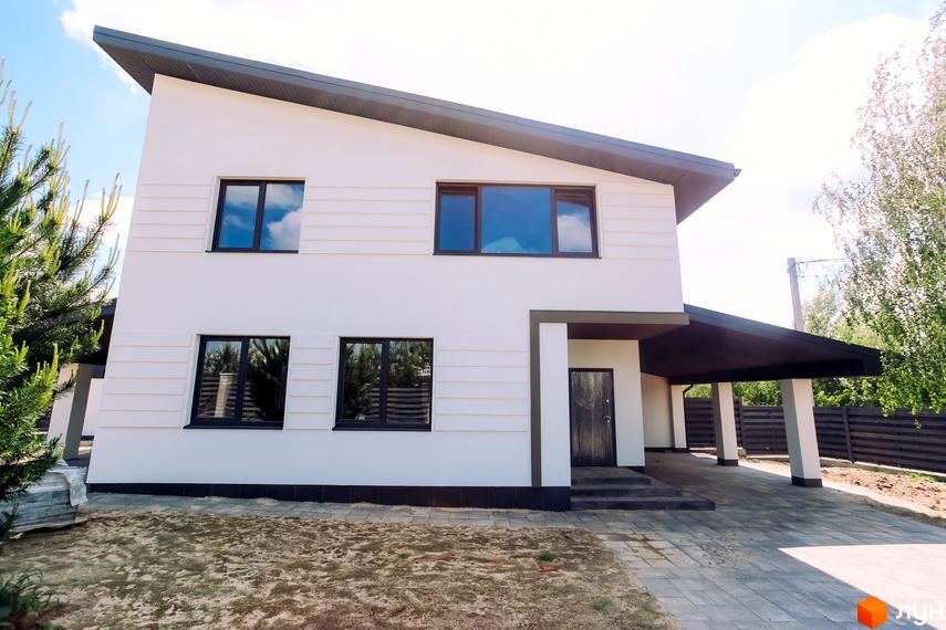 Двоповерховий будинок з білою фасадною обробкою та темним дахом. Поруч розташований навіс для автомобілів та огороджена територія.