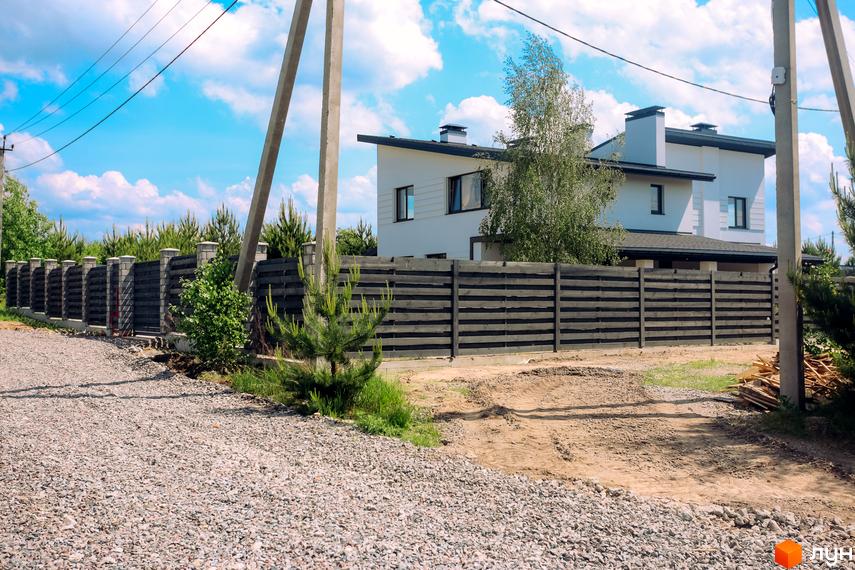 Сучасний білий дуплекс з темним дахом та вікнами, огороджений темним дерев'яним парканом. Територія біля будинку засипана гравієм та піском, є зелені насадження.