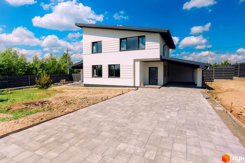 Сучасний білий двоповерховий будинок з навісом для авто та вимощеною плиткою прибудинковою територією. Зелена ділянка з парканом та деревами навколо.