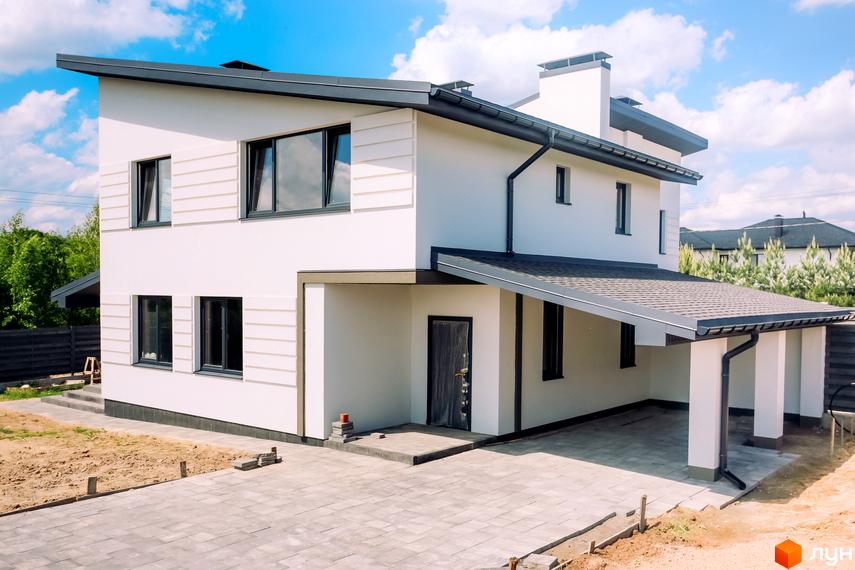 Сучасний двоповерховий будинок з білою фасадною штукатуркою та темним дахом. Біля будинку розташована прибудинкова територія з бруківкою та зеленими насадженнями.