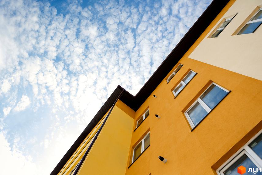 Зовнішній вигляд новобудови ЖК Сімейна Фортеця з жовтим фасадом та білими вікнами на фоні блакитного неба з хмарами. Будівельний проєкт з сучасною архітектурою.