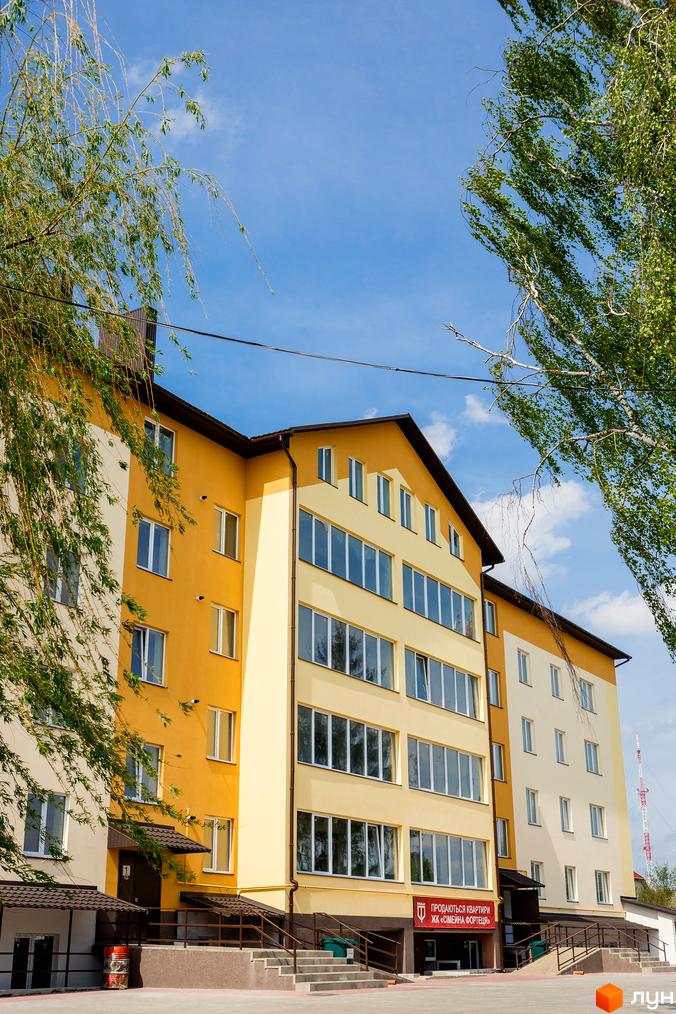 Фасад новобудови ЖК Сімейна Фортеця в жовто-бежевих тонах з великою кількістю вікон. Перед будинком розташовані сходи та навіси.