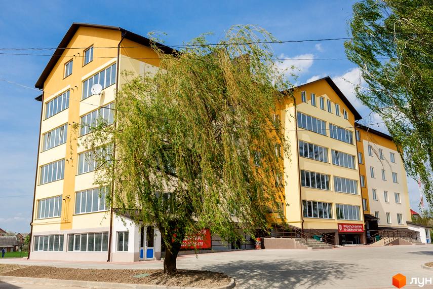 Житловий комплекс Сімейна Фортеця - жовта багатоповерхівка з великою кількістю вікон. Навколо будинку зелене дерево та облаштована прибудинкова територія.