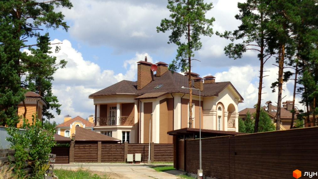 Зображення будинку в Котеджному містечку Green Wood club. Двоповерховий будинок з коричневим дахом та оздобленим фасадом, оточений деревами та зеленими насадженнями.