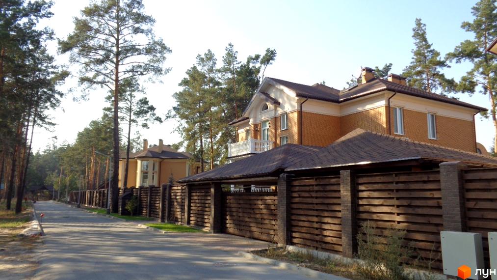 Зображення двоповерхового будинку в Котеджному містечку Green Wood club з коричневою покрівлею та дерев'яним парканом. Навколо сосновий ліс та асфальтована дорога.