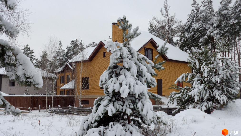 Зимовий краєвид Котеджного містечка Green Wood club: жовтий цегляний будинок з темним дахом, вкритий снігом, оточений засніженими деревами.