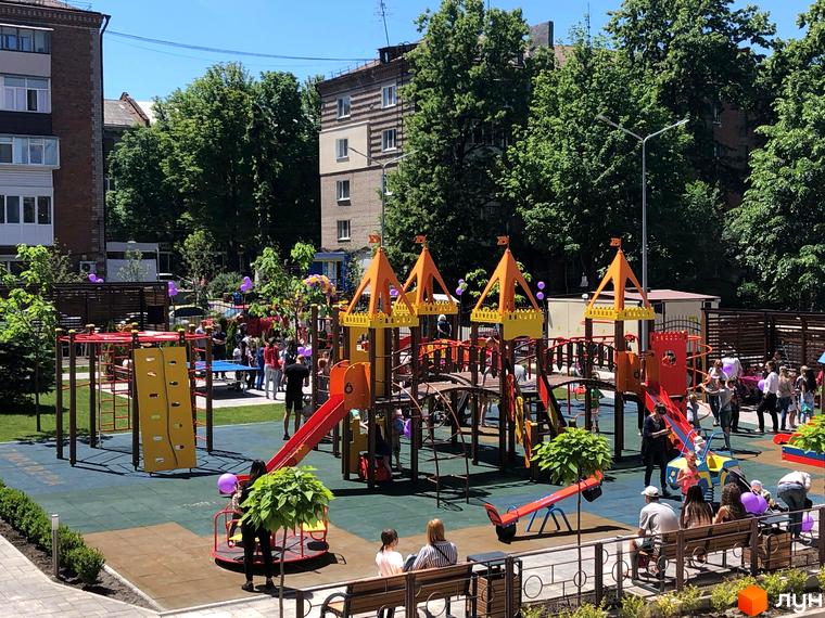 Дитячий майданчик у житловому комплексі Атмосфера з різнокольоровими гірками та гойдалками. На території багато зелені та людей.