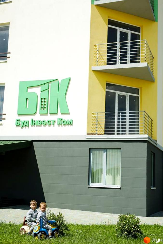 Фасад Житлового комплексу Південний з білими, жовтими стінами та балконами. На передньому плані зелений газон з дітьми, що граються.