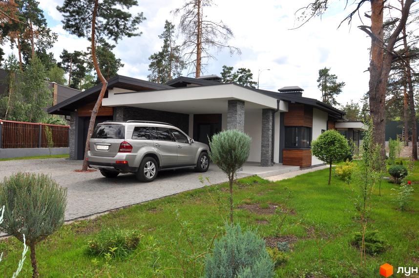 Одноповерховий будинок з сірим фасадом та критим паркінгом. На території розташовані дерева та газон.