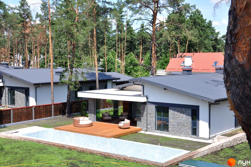 Будинки Котеджного містечка Хвойний квартал з білими стінами, сірими дахами та кам'яним облицюванням. На подвір'ї басейн, дерев'яна тераса та хвойні дерева.