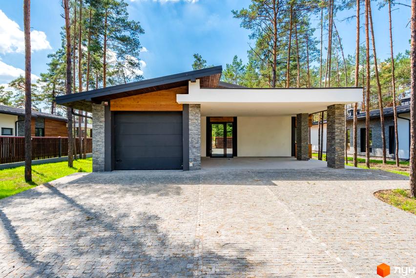 Будинок Котеджного містечка Хвойний квартал з темною гаражною брамою та критим паркувальним місцем. Територія має бруківку та хвойні дерева.